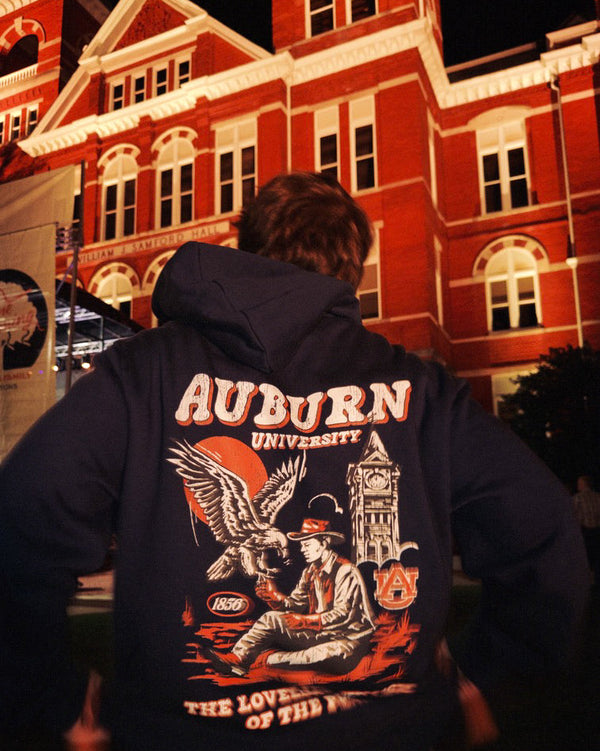 A person viewed from behind wearing a navy blue hoodie with a large Auburn University graphic on the back, standing in front of a warmly lit red brick building at night.
