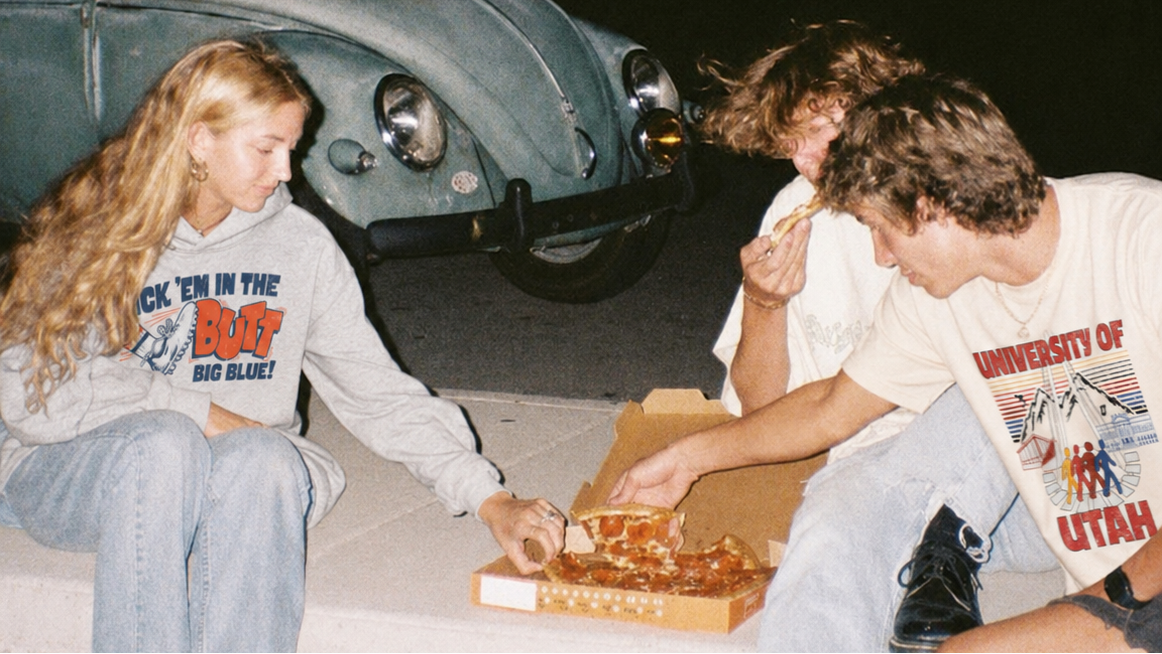 Three young adults sitting on a concrete ledge at night, sharing a pizza box while wearing collegiate apparel from Saturday Co.